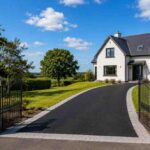 Newly installed black SMA tarmac driveway with silver-grey block paving borders leading through open wrought-iron gates to a detached home in Castlepollard, County Westmeath, with green lawns, mature trees and rolling countryside under a bright summer sky.
