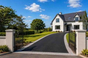 Newly installed black SMA tarmac driveway with silver-grey block paving borders leading through open wrought-iron gates to a detached home in Castlepollard, County Westmeath, with green lawns, mature trees and rolling countryside under a bright summer sky.